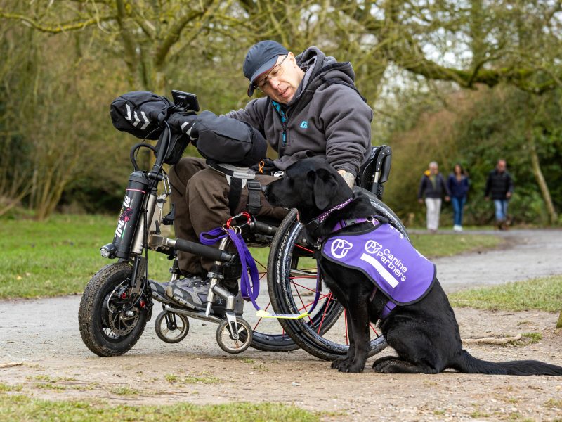 James in black wheelchair with canine partner Dawn sat next to him in purple jacket outside