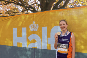 A woman with blonde hair is holding her medal in front of a half marathon sign. She is wearing a Canine Partners running vest.
